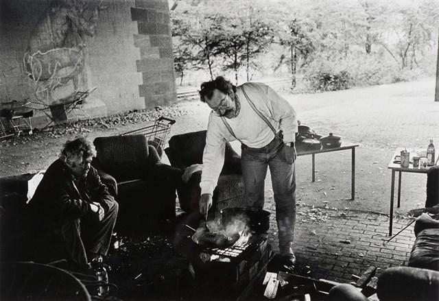 Das Schwarz-Weiß-Foto von Absiag Tüllmann zeigt eine Gruppe Obdachloser die im Freien auf Sesseln um einen Grill sitzen und stehen