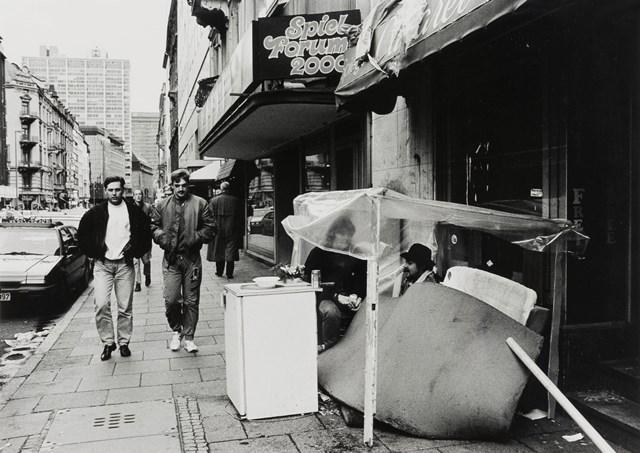 Das Schwarz-Weiß-Foto von Absiag Tüllmann zeigt einen Bürgersteig neben einer Straße, zwei Männer laufen auf die Kamera zu, auf der rechten Bildseite sitzen zwei Obdachlose unter einer Plastikplane.