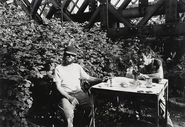 Das Schwarz-Weiß-Foto von Abisag Tüllmann zeigt zwei Obdachlose Personen, die an einem Tisch vor einem Gebüsch sitzen.