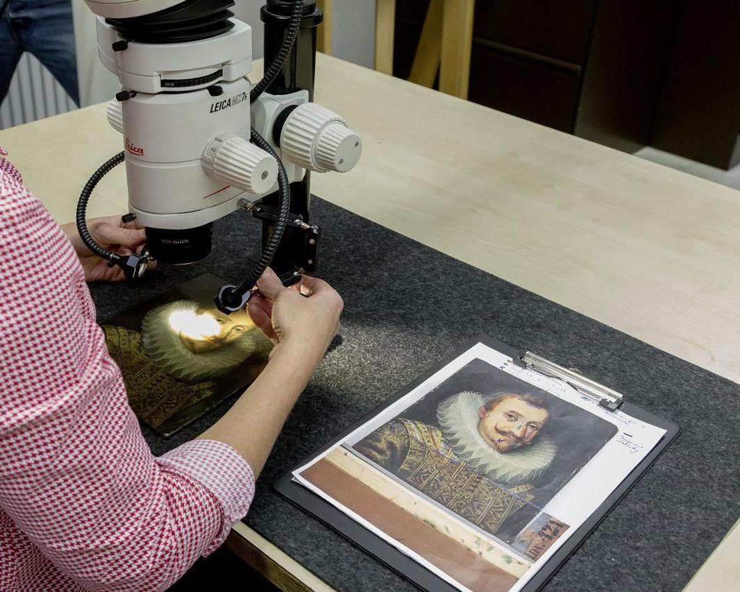 Das Foto zeigt eine Person an einem Mikroskop, unter dem ein Gemälde liegt. Daneben liegen Notizen. 
