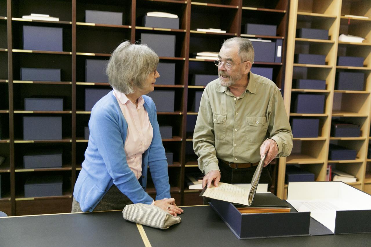 Zwei Personen stehen in der Bibliothek der Generationen vor einem Regal mit Kassetten. Geöffnet vor ihnen liegt eine geöffnete Kassette mit Mappen und Schriftstücken.