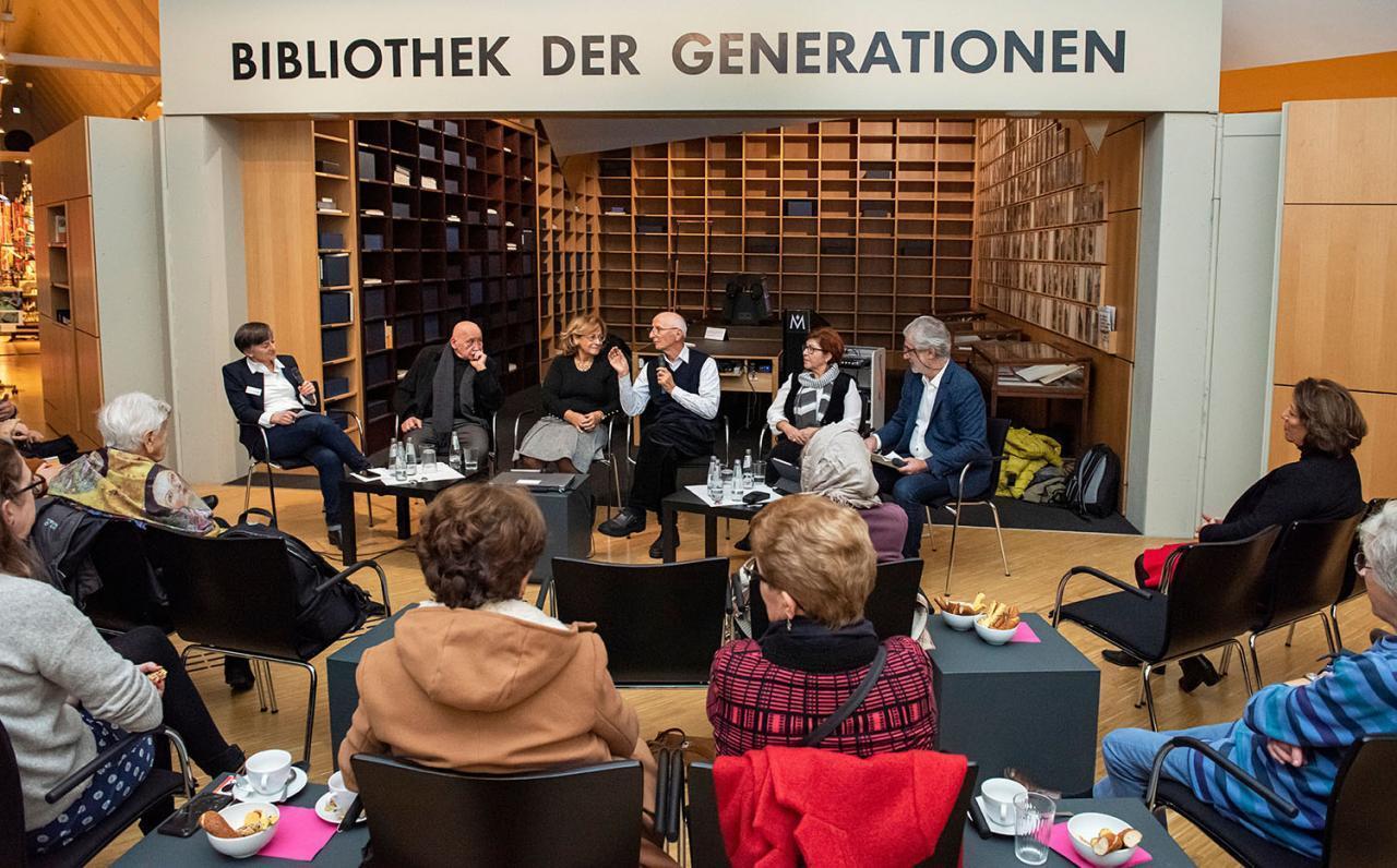 Vor der Regalinstallation der Bibliothek der Generationen sitzen sechs Personen, deren Gespräch ein Publikum zuhört.