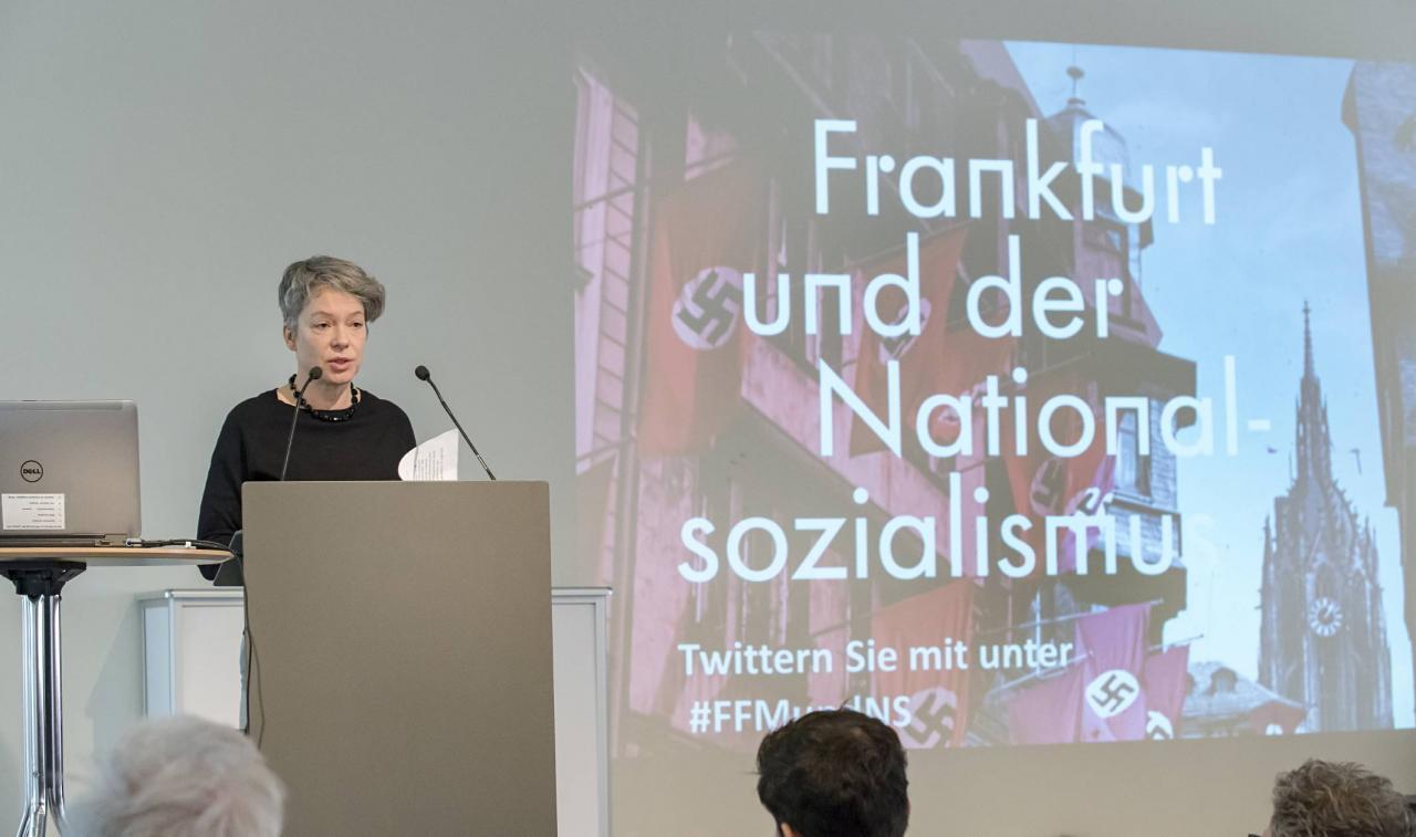 Das Foto zeigt Kulturdezernentin Dr. Ina Hartwig bei ihrem Grußwort, im Hintergrund ist das Veranstaltungsplakat zu sehen.