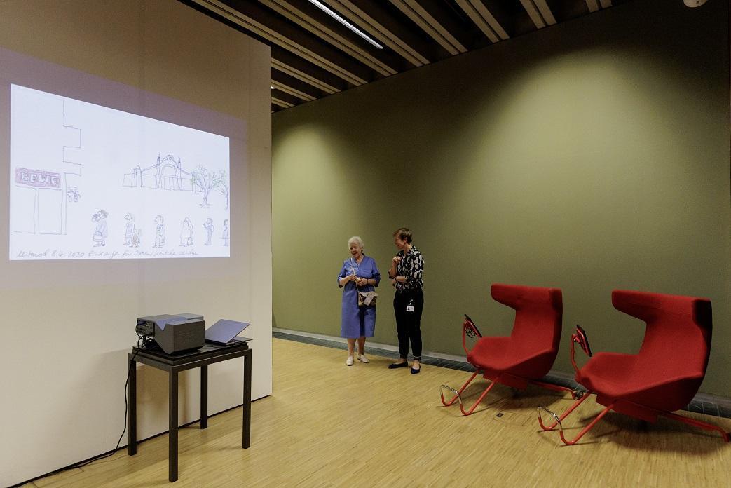 Im Ausstellungsraum wird eine Zeichnung an die Wand projiziert. Zwei rote Sessel mit Tablet sind im Hintergrund zu sehen. Zwei Menschen stehen daneben und unterhalten sich.