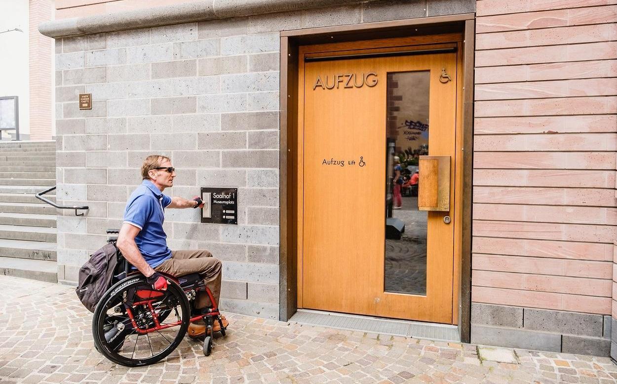 Das Foto zeigt einen Rollstuhlfahrer, der an der Tür zum Aufzug läutet. 
