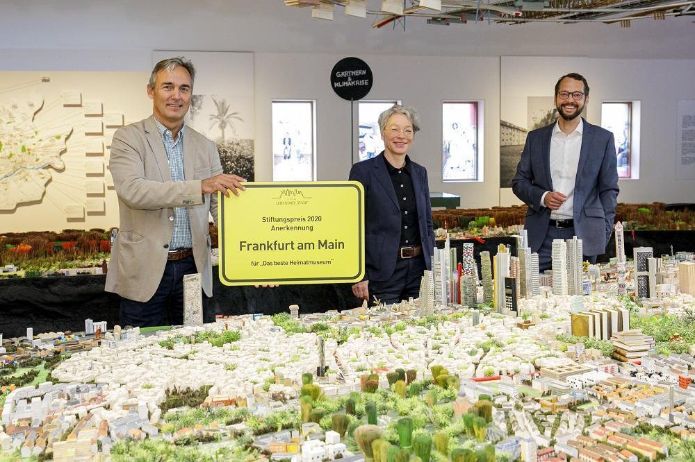 Das Foto zeigt drei Personen (Museumsdirektor Jan Gerchow, Kulturdezernentin Ina Hartwig und Jan Schneider, Stiftung Lebendige Stadt, vor dem Stadtmodell. Herr Gerchow hält ein gelbes Schild mit schwarzem Text: Stiftungspreis 2020 Anerkennung Frankfurt am Main für das beste Heimatmuseum. 