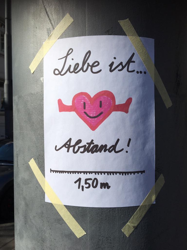 Das Foto zeigt einen Zettel an einem Laternenmast, auf dem "Liebe ist Abstand" geschrieben steht, außerdem ist ein Herz mit ausgestreckten Armen gezeichnet. 