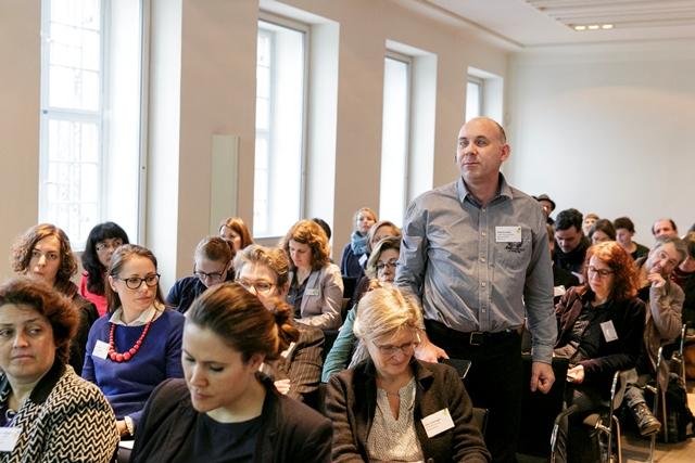 Die Fotografie zeigt das sitzende Publikum der Fachkonferenz. Ein Mann steht in der Mitte des Publikums und sagt etwas.